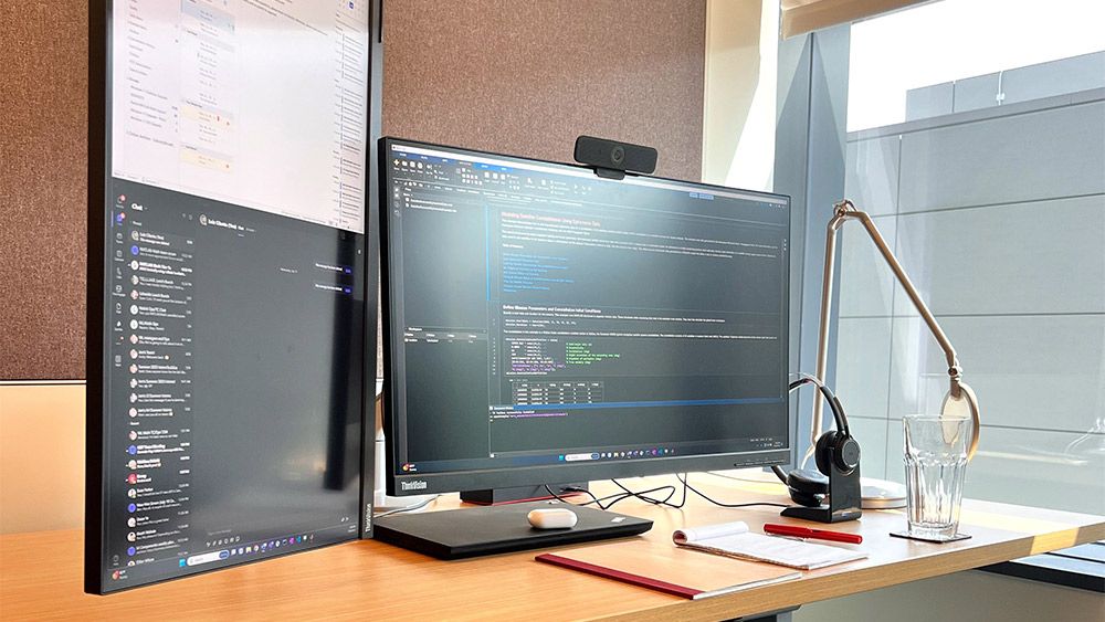 Height-adjustable desk with two large monitors, laptop, and headset. One monitor shows Outlook and Teams, and the other shows MATLAB and the team’s case management software.