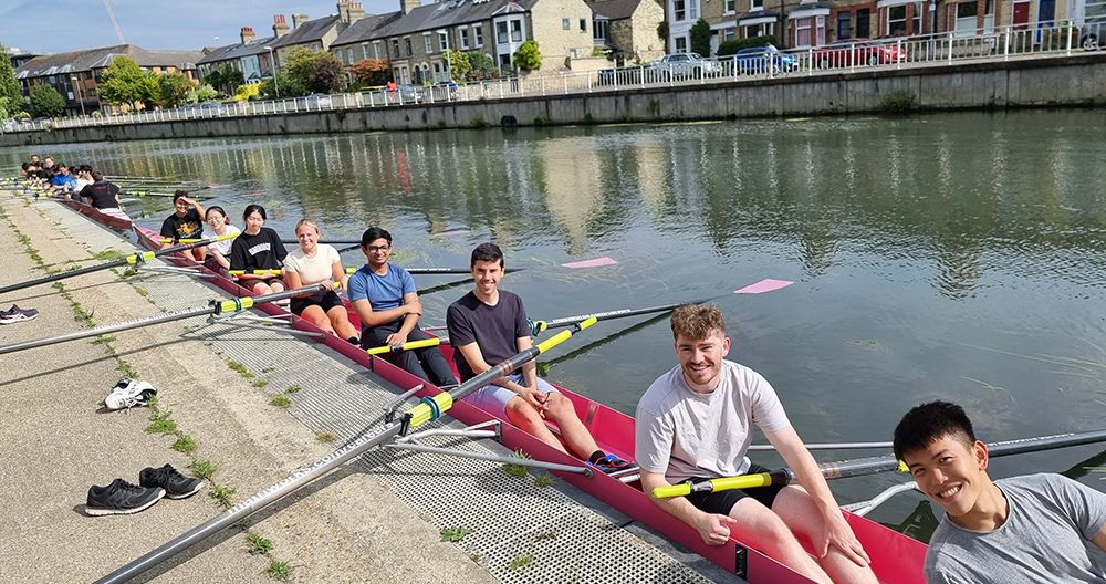 A group of people sitting in a long rowing boat on a calm river, smiling at the camera, with houses in the background.
