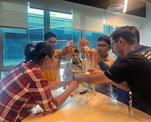 Several people collaborate to build a structure using spaghetti and marshmallows on a table in a bright room.