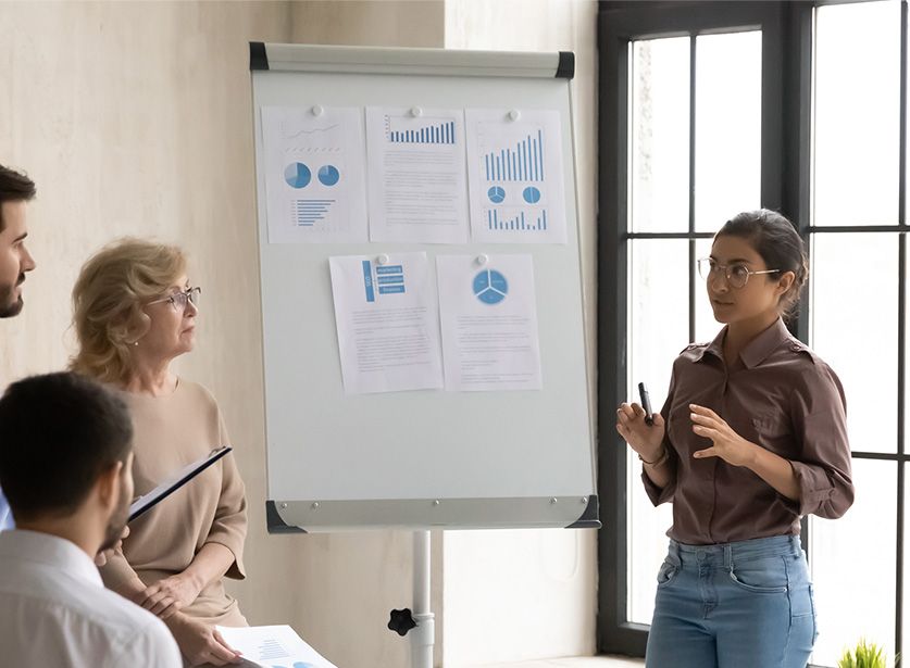 A woman presents charts and graphs on a flip chart to a small group during a meeting in a bright room.