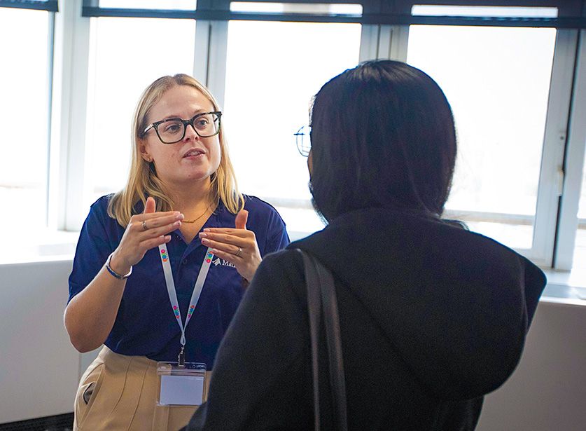 Two women have a discussion indoors; one gestures while speaking, wearing a lanyard, and the other listens attentively.