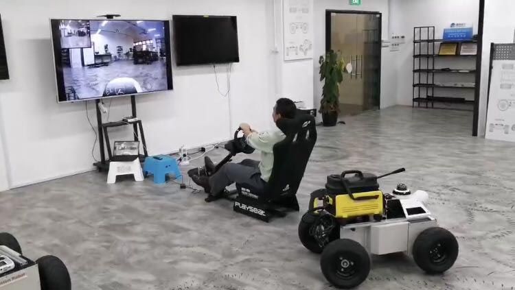 The robot is shown in the foreground. An operator is seated in front of a large screen that shows the robot’s surroundings.  The operator is controlling the robot with a steering wheel. 