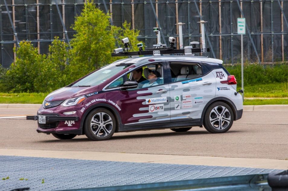Two people sitting in the Texas A and M autonomous vehicle, which is parked in a lot. The car has purple and white stripes with the names of sponsors on the car.